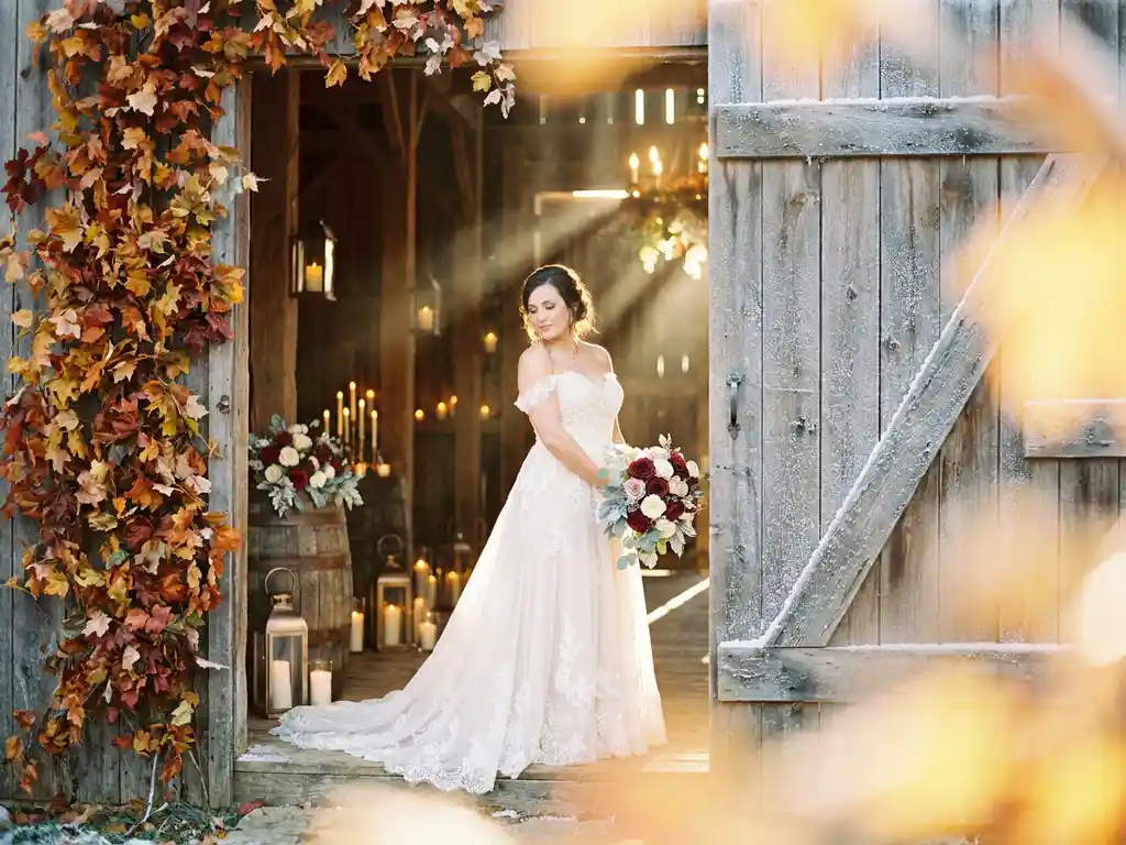 Braut im weißen Hochzeitskleid in rustikaler Scheunentür, herbstliches Laub und Winterfrost, warmes Kerzenlicht, goldenes Gegenlicht.
